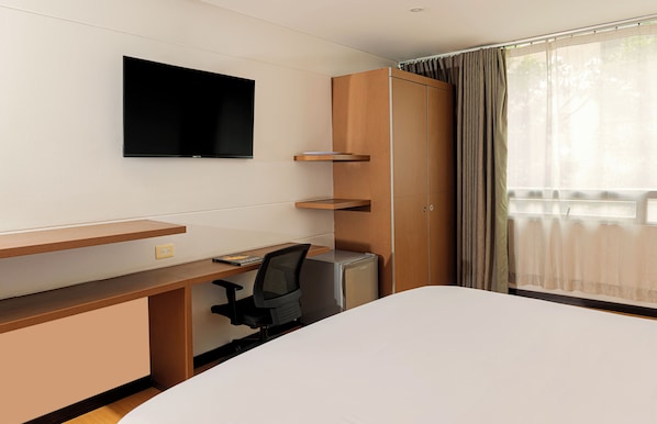 A hotel room with a bed, a desk with a computer, a TV mounted on the wall, and a view of the outdoors through the window.