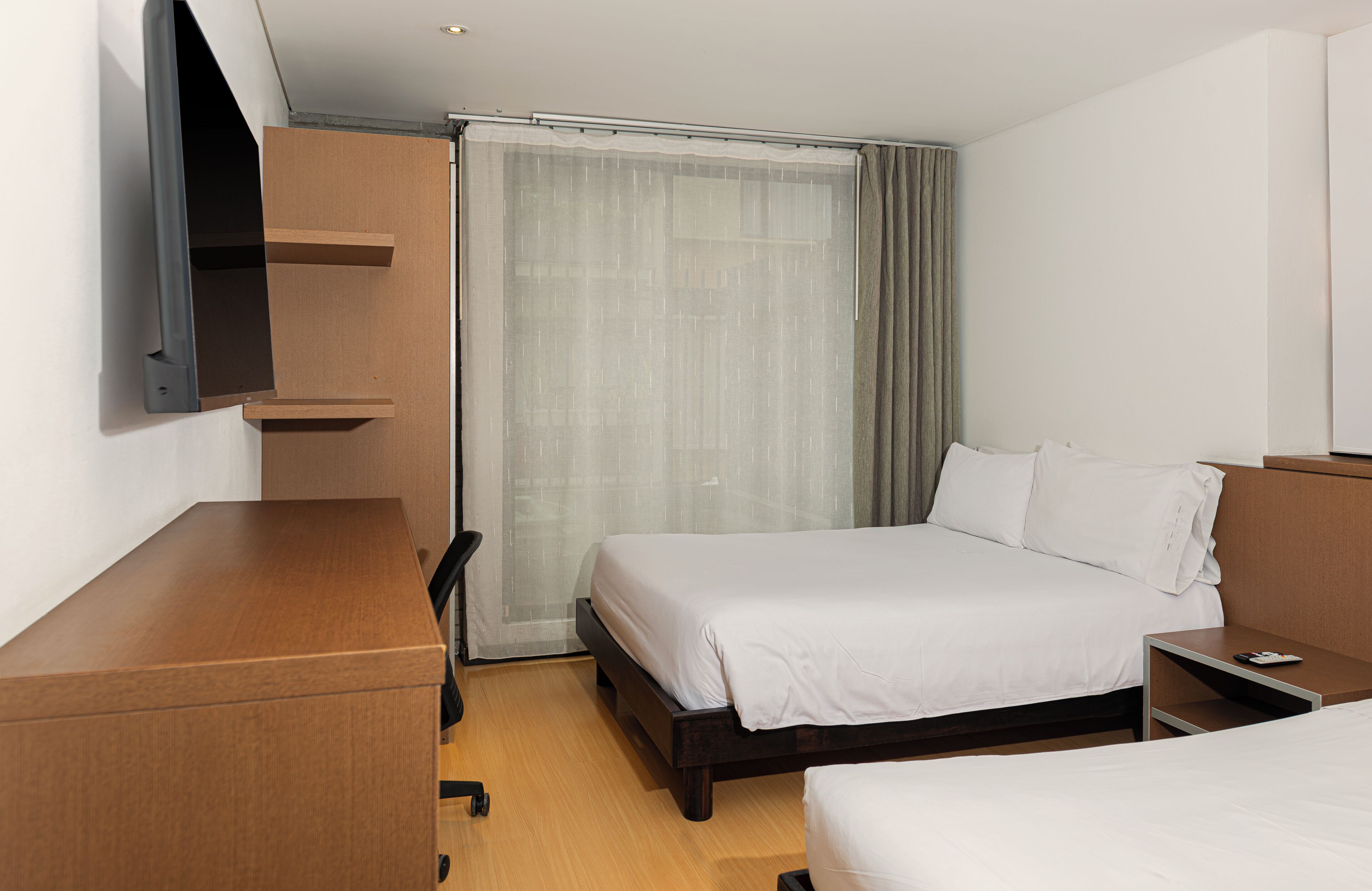 Habitación de hotel con cama, escritorio, silla, televisión montada en la pared y ventana con cortinas.