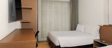Habitación de hotel con cama, escritorio, silla, televisión montada en la pared y ventana con cortinas.