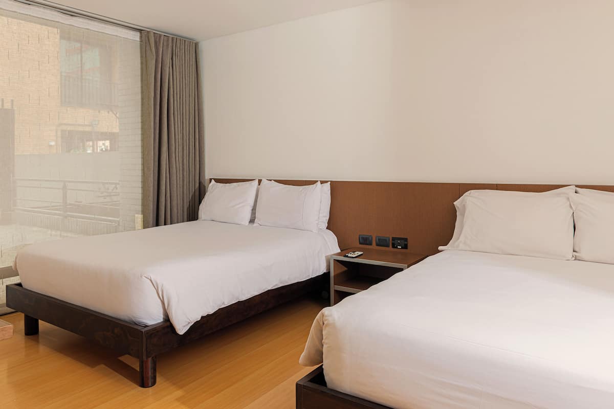 Quarto de hotel com duas camas, uma cabeceira de madeira, mesa de cabeceira e janela com vista para um edifício de tijolos.