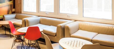 Een moderne lounge met leren banken, rode en witte stoelen en tafels. Grote ramen zorgen voor natuurlijk licht en uitzicht op de omgeving.