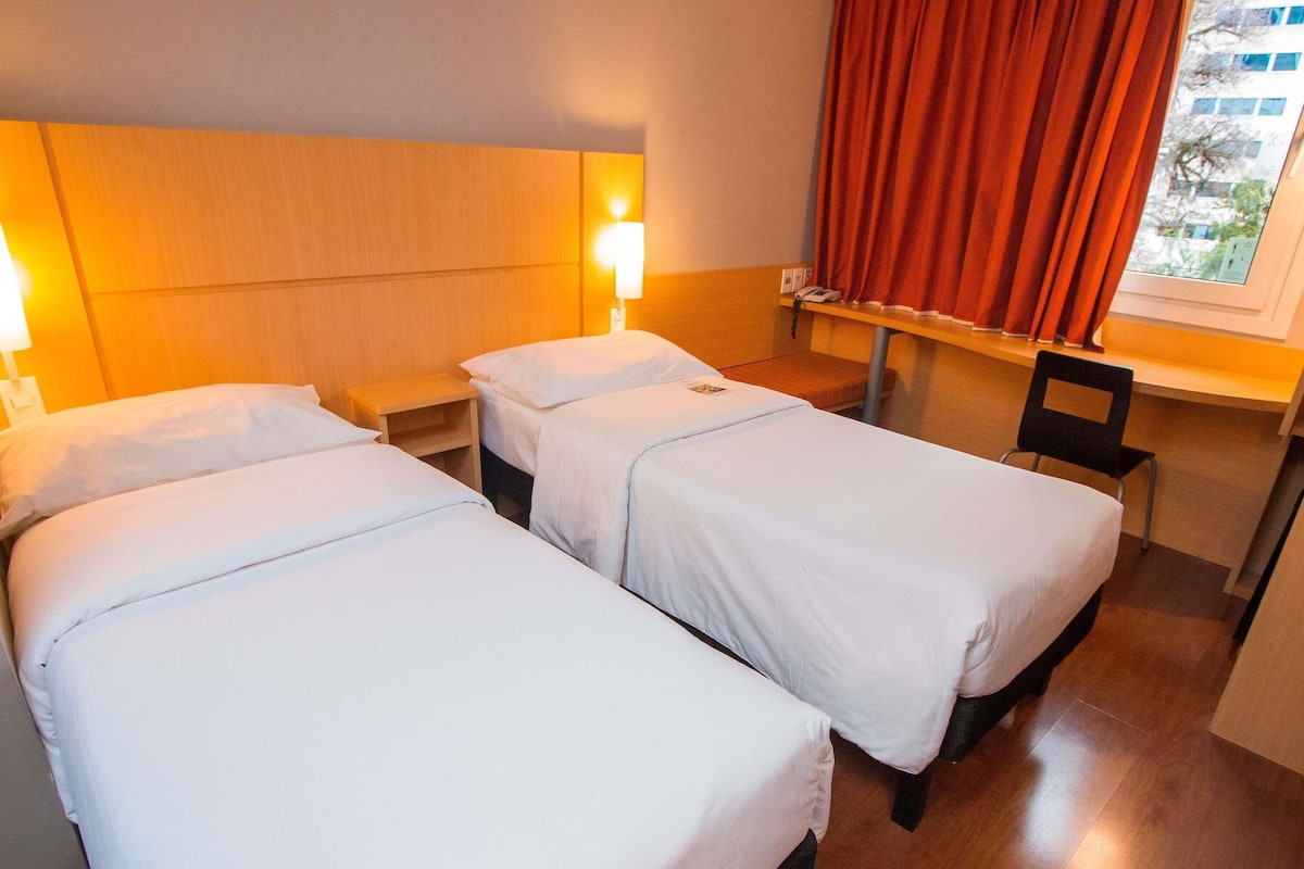 Habitación de hotel con dos camas individuales, una mesita de noche, una silla, un ventanal con cortinas rojas y vistas a edificios.