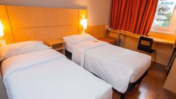 Habitación de hotel con dos camas individuales, una mesita de noche, una silla, un ventanal con cortinas rojas y vistas a edificios.