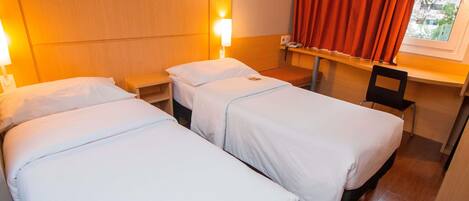 A hotel room with two single beds, a nightstand, a chair, a window with red curtains, and a view of buildings outside.