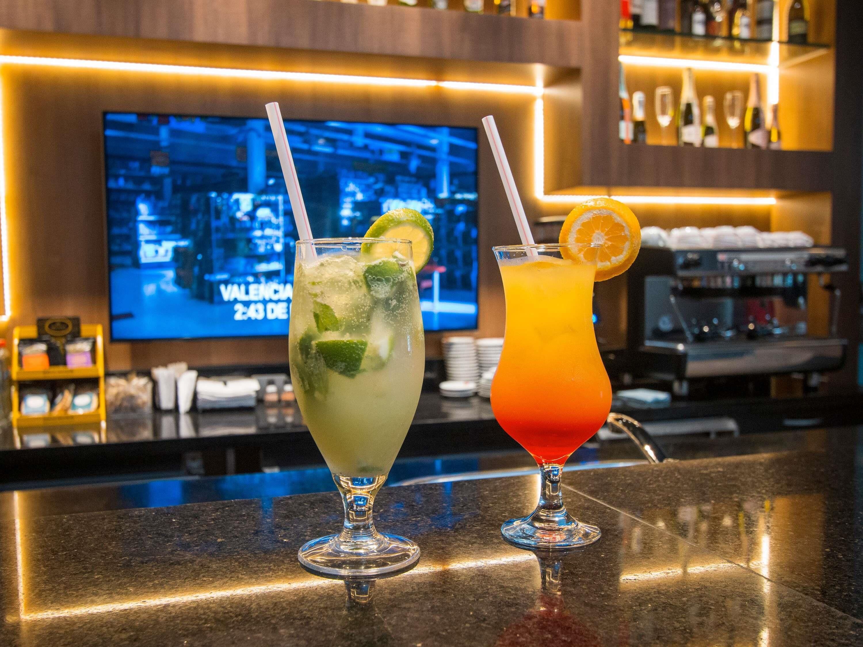 Two cocktails on a bar counter with a TV displaying 'VALENCIA 2:43 DE' in the background.