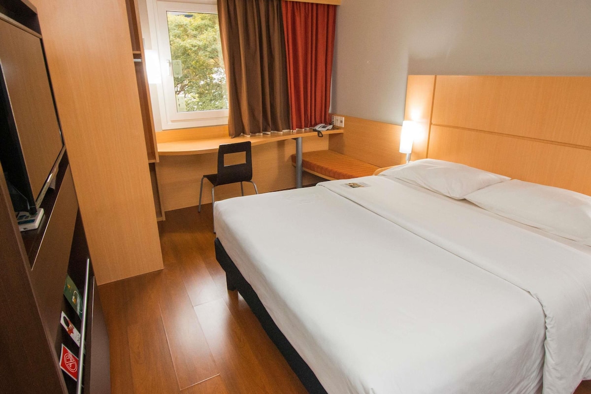 Una habitación de hotel con cama, escritorio, silla y ventana con cortinas.