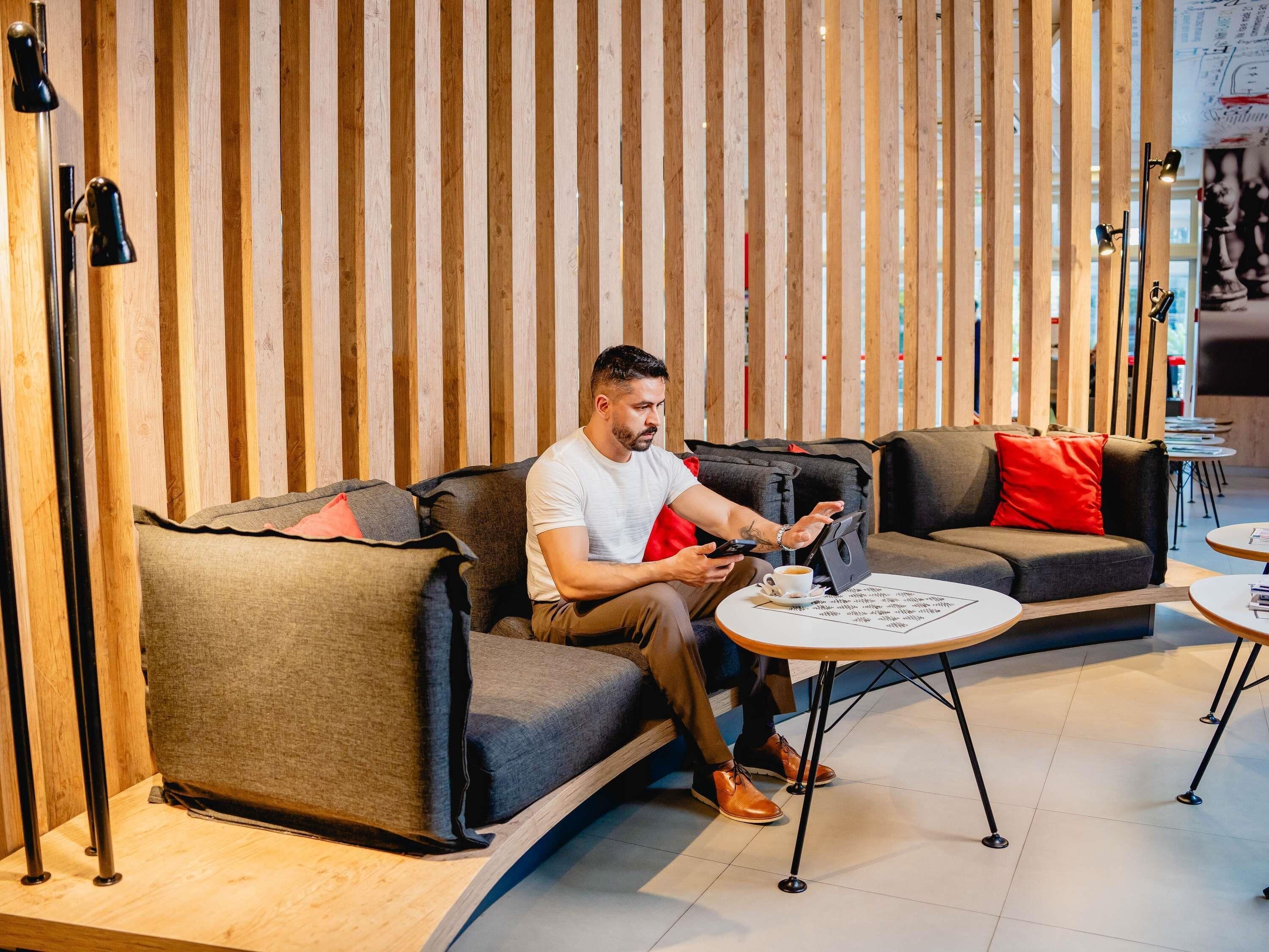 A man sitting on a sofa in a modern interior with a wooden slat wall, a small table with a cup, and a tablet.