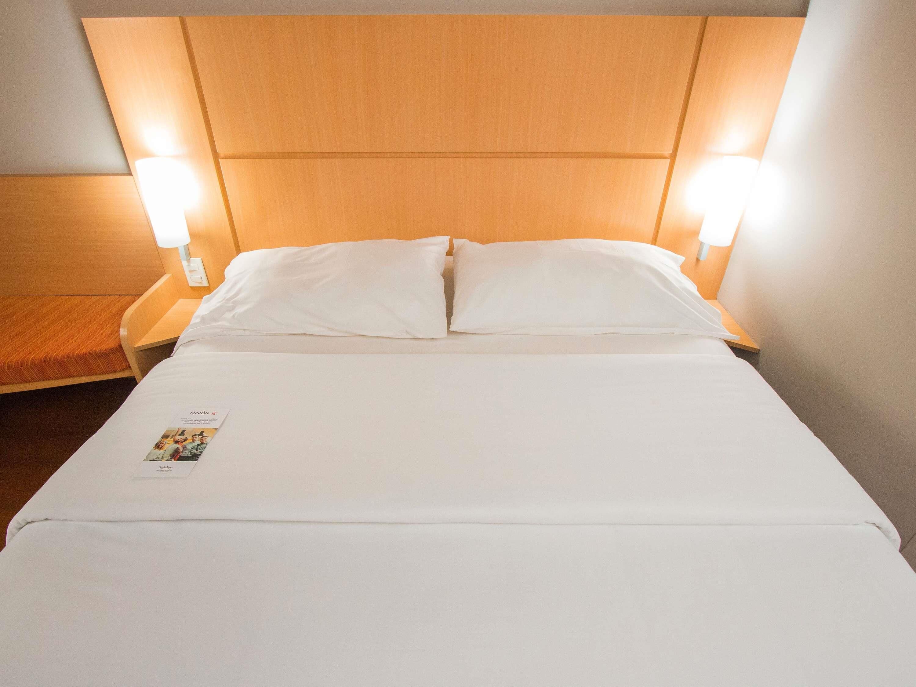 Cama bem arrumada com lençóis brancos e duas luminárias de cabeceira.