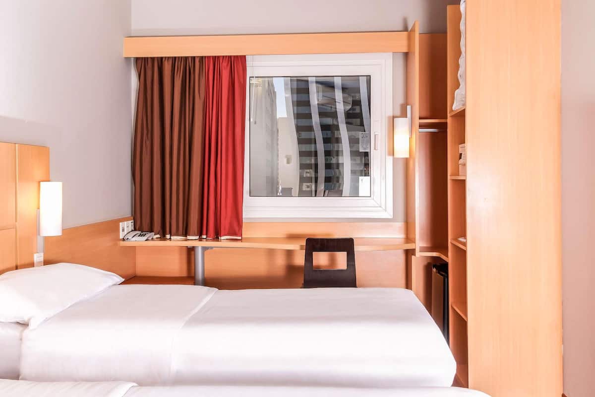 A hotel room with a bed, a desk, a chair, a window with curtains, and a bedside lamp.