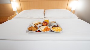 Un lit bien fait, avec du linge de lit blanc et des oreillers, un plateau de petit-déjeuner comprenant des œufs, des fruits et des viennoiseries, et deux lampes de chevet.