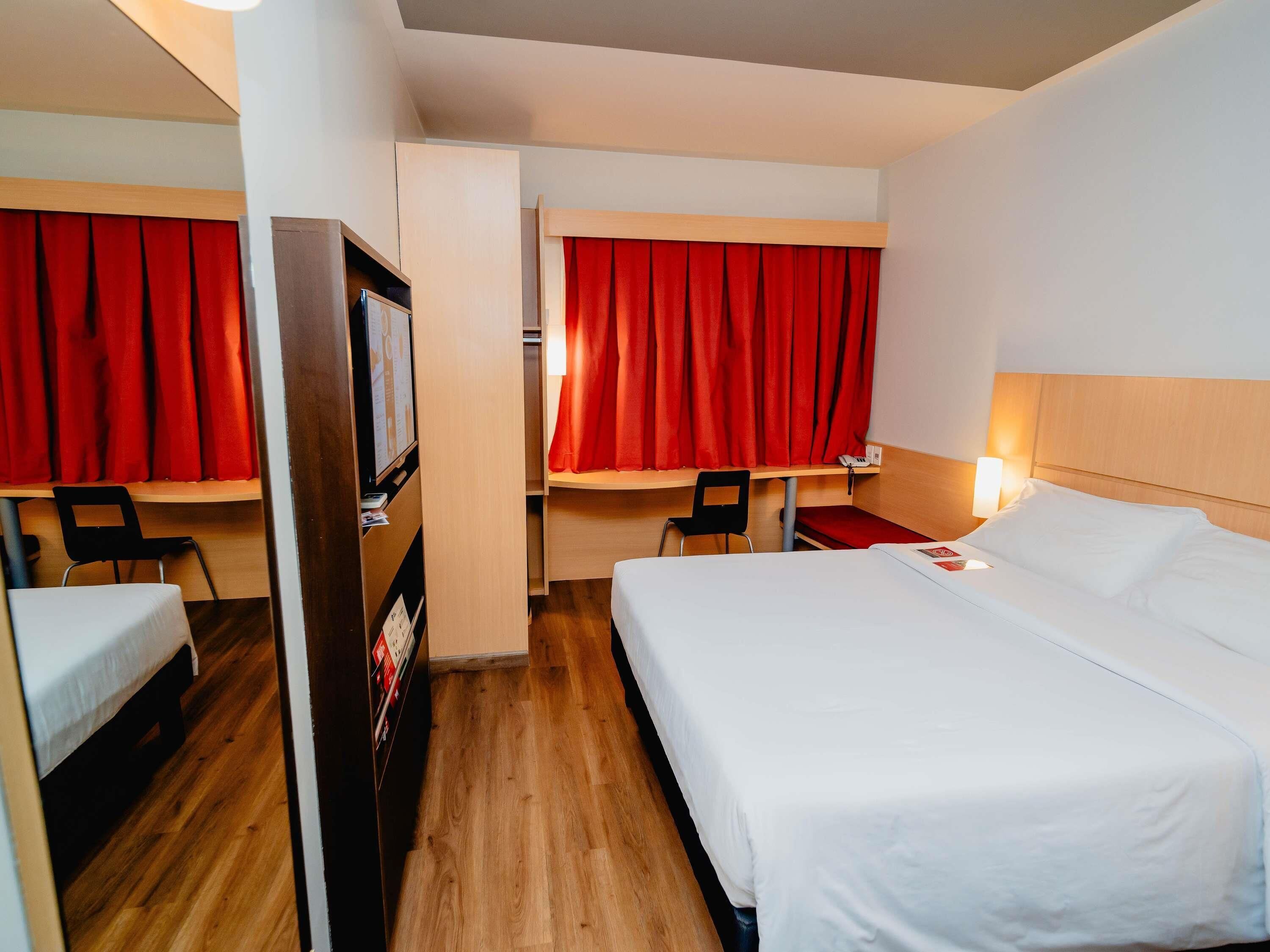 A hotel room with a bed, a desk, a chair, a TV, and red curtains.