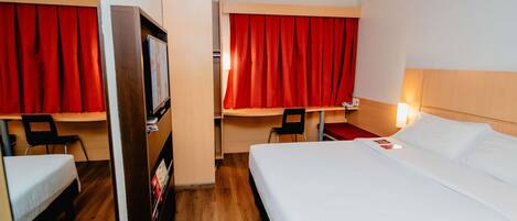 A hotel room with a bed, a desk, a chair, a TV, and red curtains.