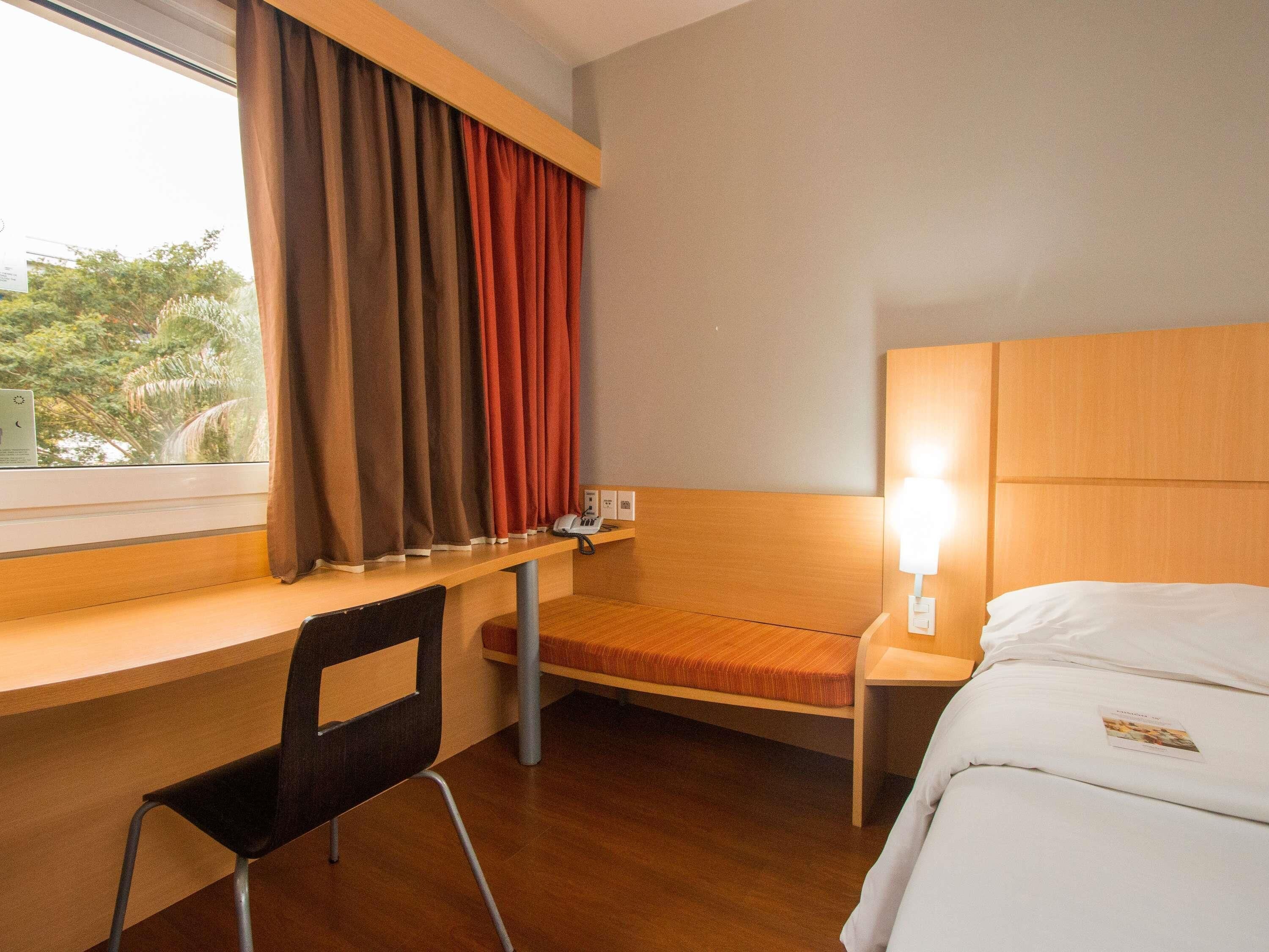 A hotel room with a bed, a desk with a chair, a nightstand with a lamp, and a window with curtains.