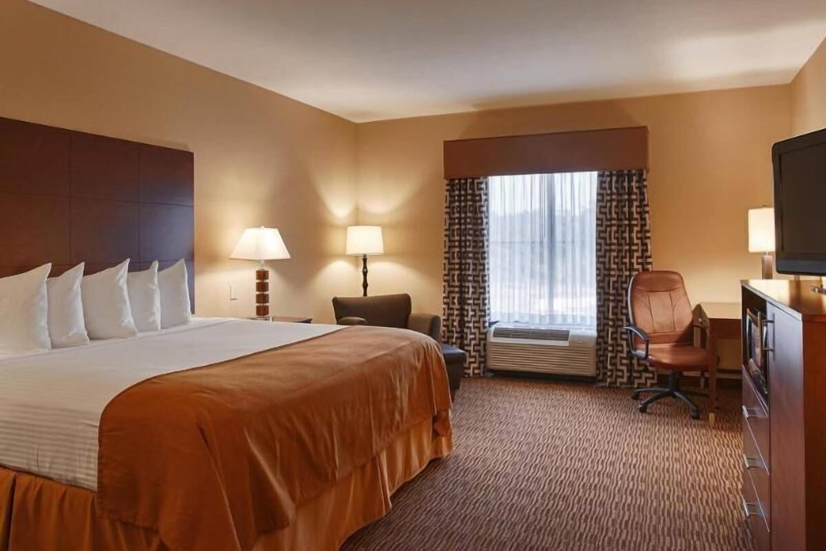 A hotel room with a large bed, two bedside lamps, a desk with a chair, a television, and an air conditioning unit.