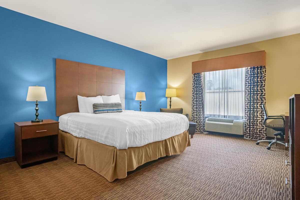 A hotel room with a large bed, two bedside tables with lamps, a chair, a desk, and a window with patterned curtains.