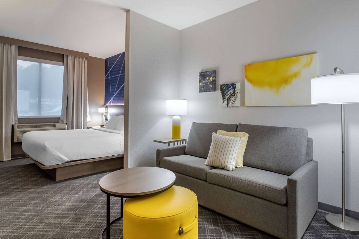 A hotel room with a bed, a sofa, a small round table, a yellow ottoman, and a lamp.