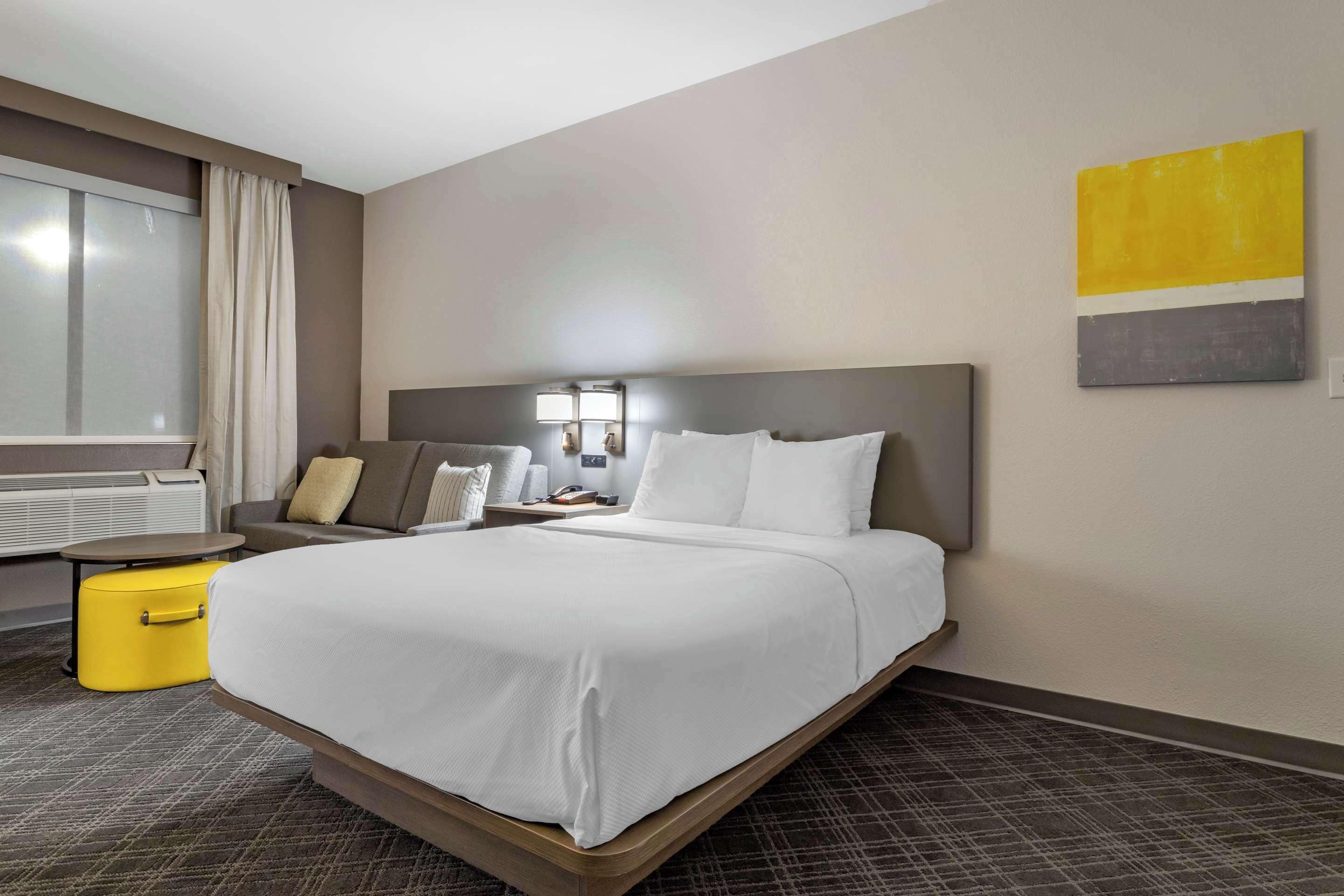 A hotel room with a large bed, a bedside table with a lamp, a small yellow ottoman, and a window with curtains.