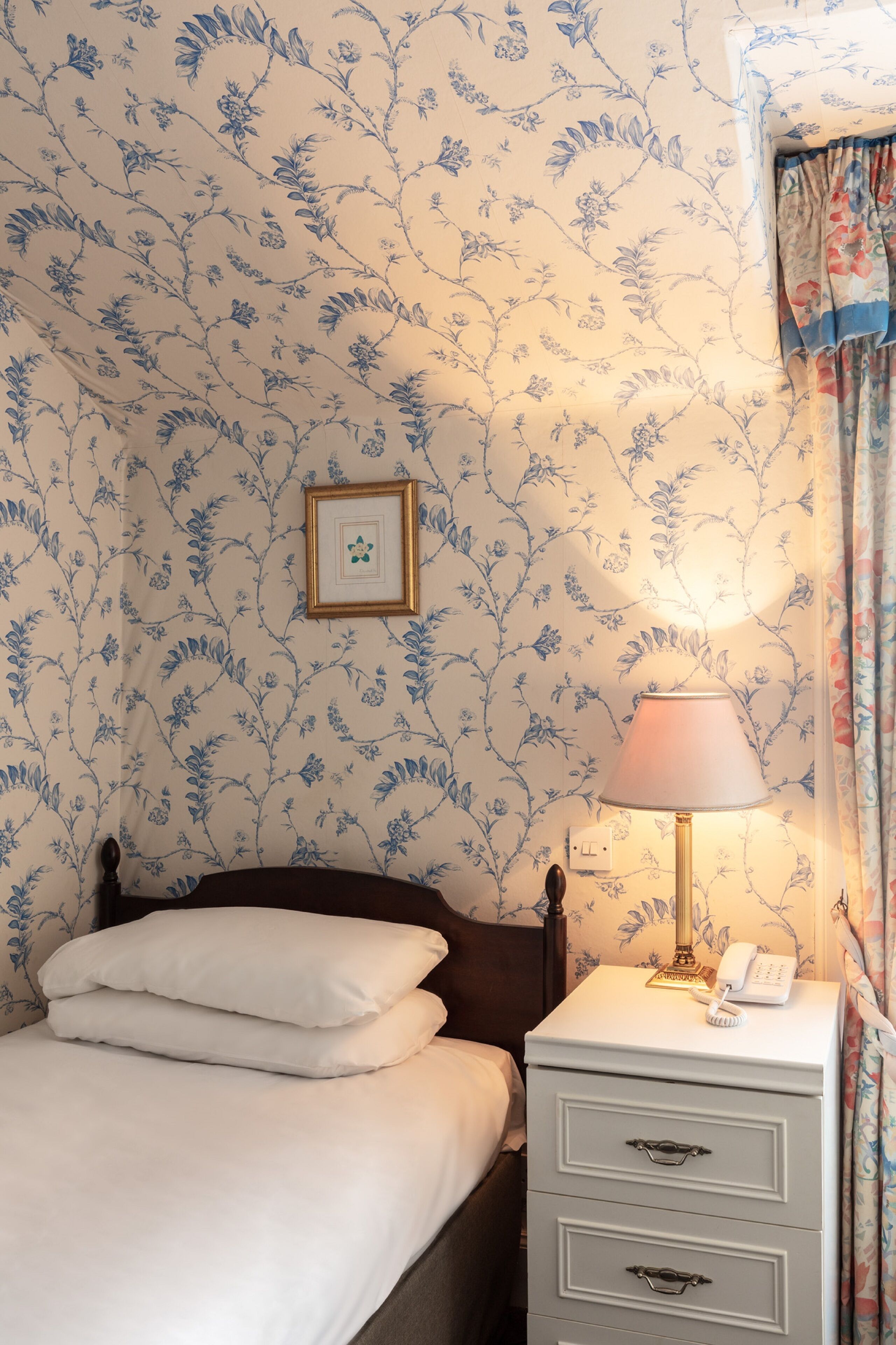 A neatly made bed with white linens, a bedside lamp, and a small bedside table with a phone and a picture frame.