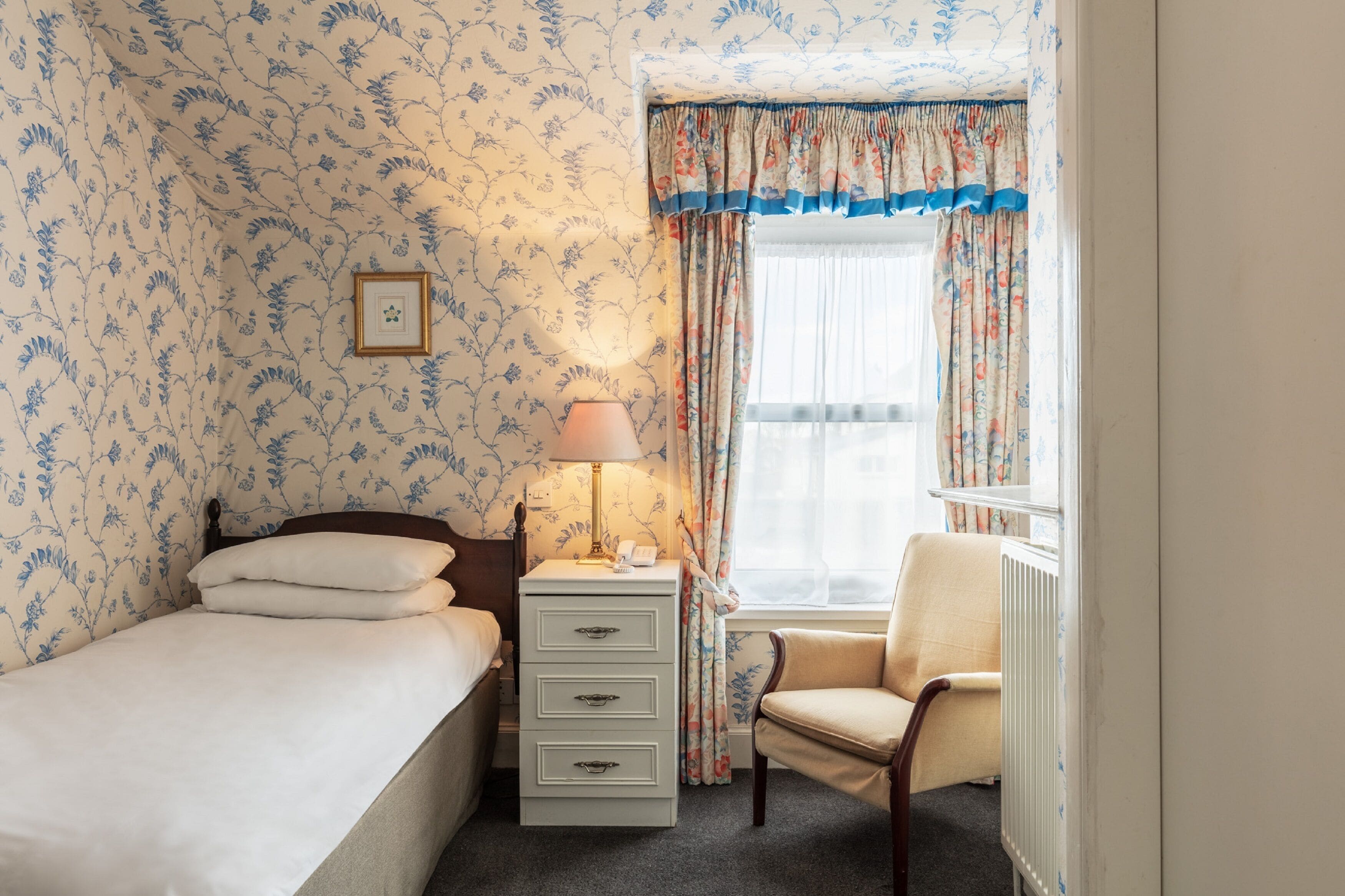 Een slaapkamer met bloemenbehang, een bed, een nachtkastje met een lamp, een stoel en een raam met gordijnen.