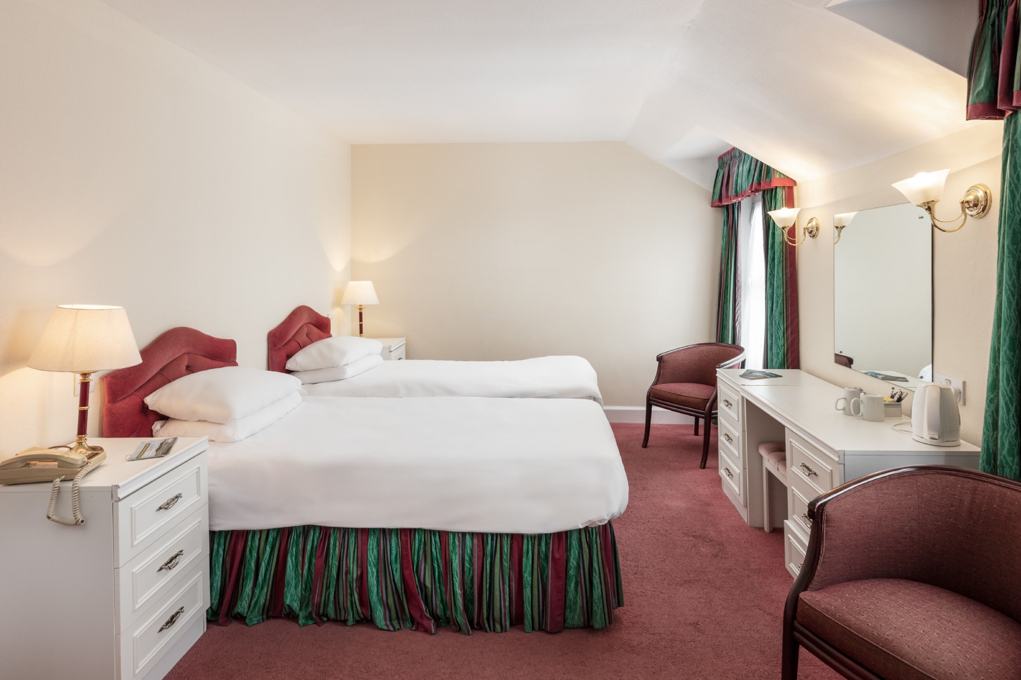 A hotel room with two beds, a desk, a chair, a dresser, a mirror, and a lamp.