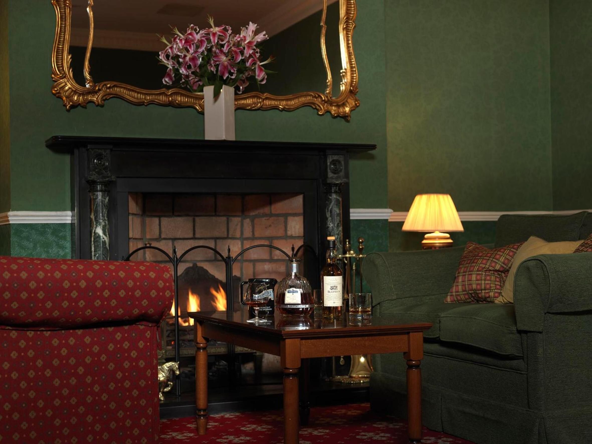 A room with a fireplace, a table with bottles, a sofa, and a red patterned armchair.