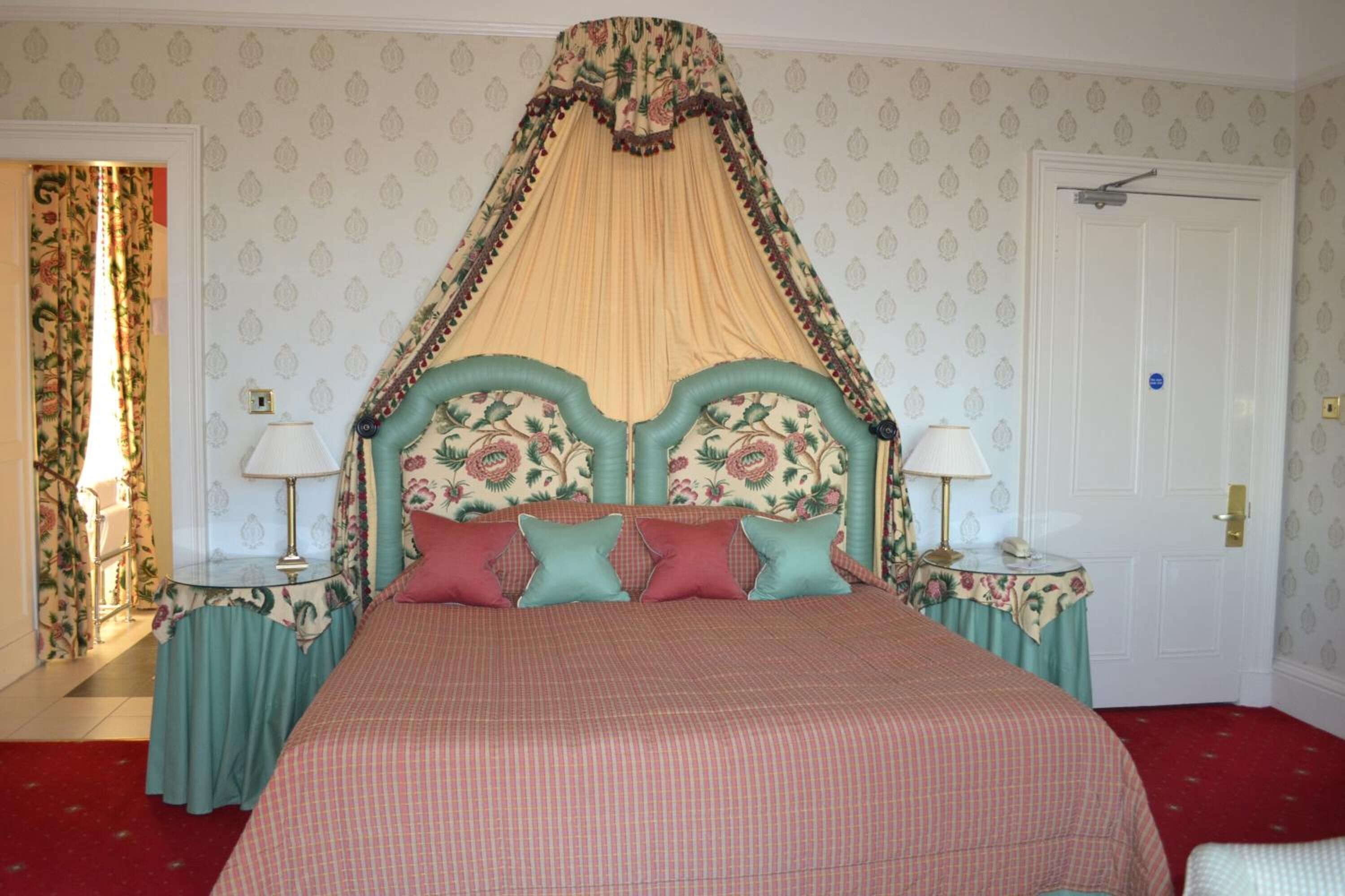 Uma cama de dossel com cabeceira e cortina floral, duas mesas de cabeceira com abajures e papel de parede estampado.