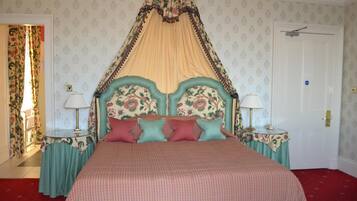 Uma cama de dossel com cabeceira e cortina floral, duas mesas de cabeceira com abajures e papel de parede estampado.