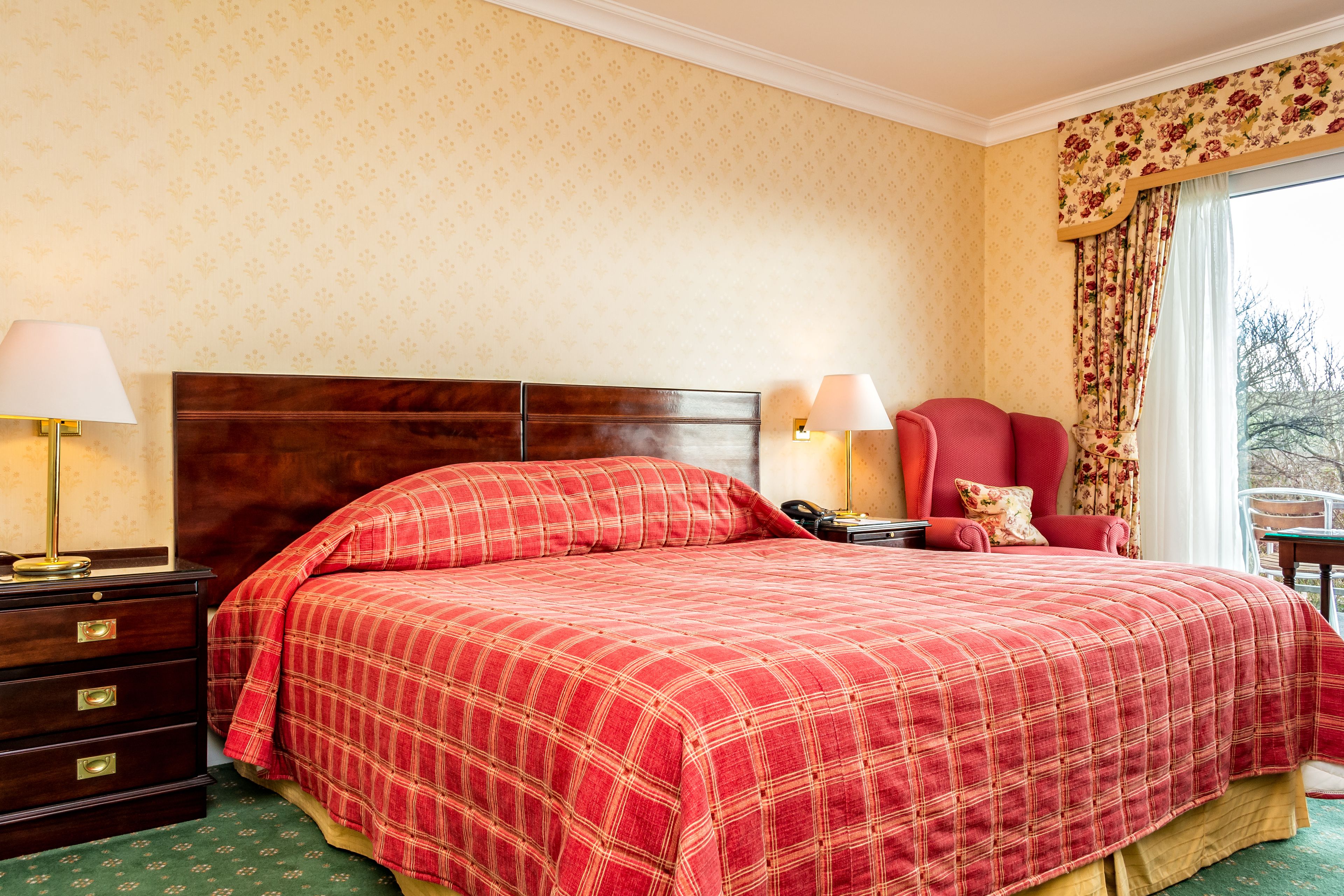 Habitación de hotel con una cama grande, dos mesitas de noche con lámparas, un sillón rojo y vistas al exterior a través de una ventana con cortinas florales.
