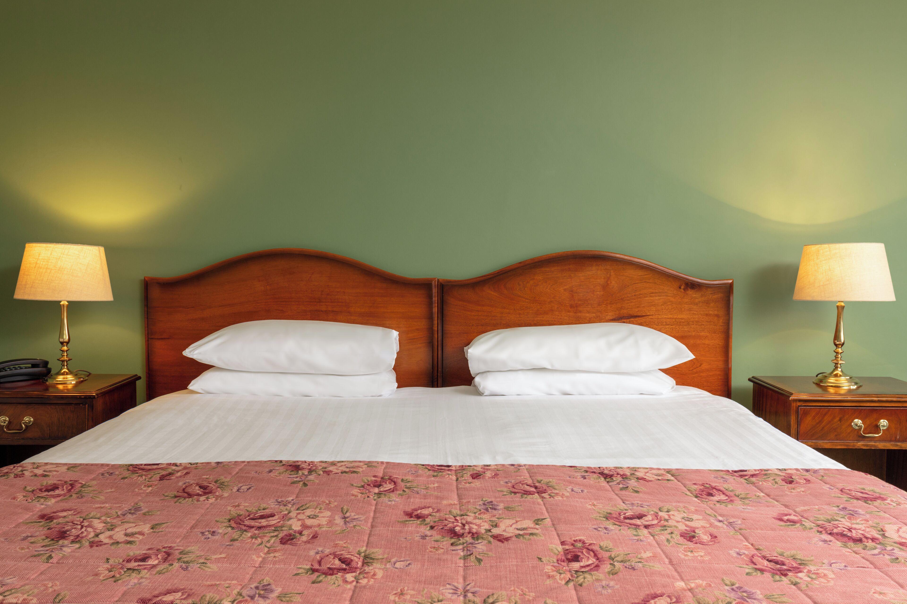 Ein Doppelbett mit floralem Überwurf, zwei weiße Kissen und ein hölzernes Kopfteil. An beiden Seiten des Bettes stehen jeweils Nachttische mit Lampen.