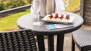 A table with a bottle, two wine glasses, and a plate of desserts on a balcony overlooking a marina and buildings. - Fernhill Hotel (Stranraer)