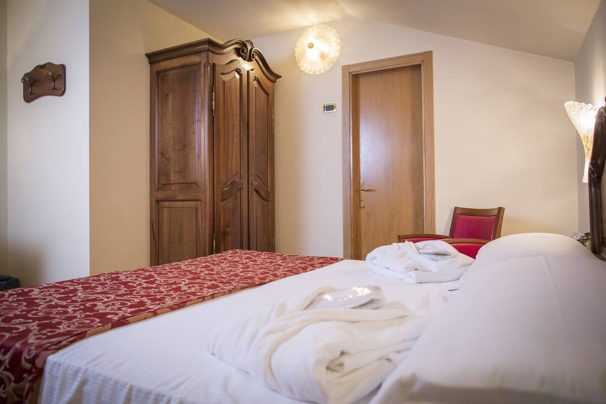 A hotel room with a bed, a wooden wardrobe, a red and white patterned bedspread, and a red chair.