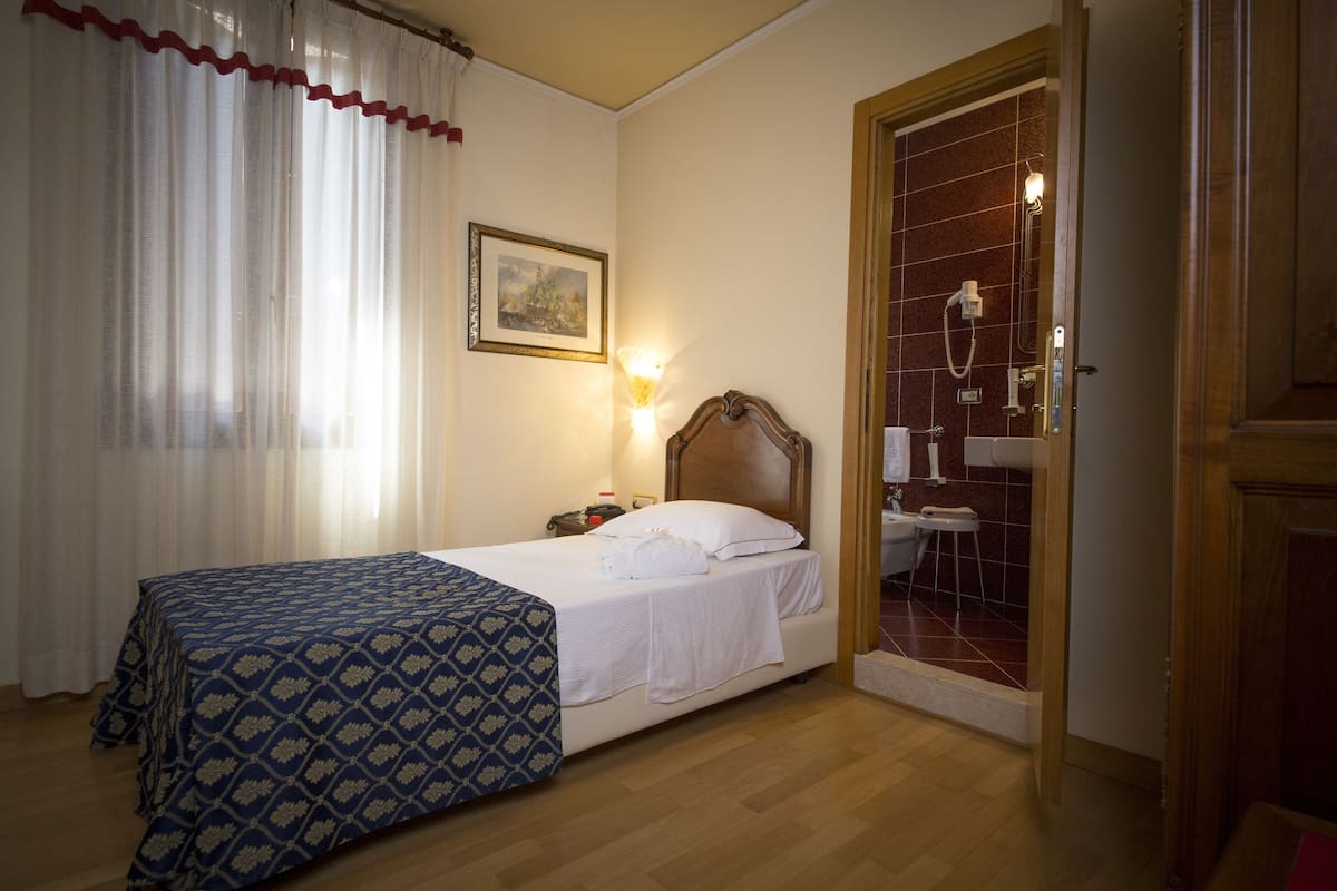 A hotel room with a bed, a bedside table, a lamp, a painting on the wall, and a bathroom visible through an open door.