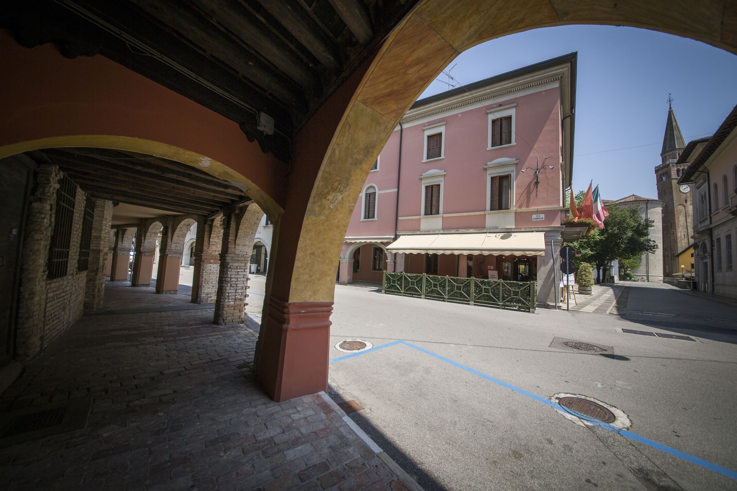 An outdoor corridor with stone arches, a paved street, and buildings with a pink facade and a clock tower in the background.