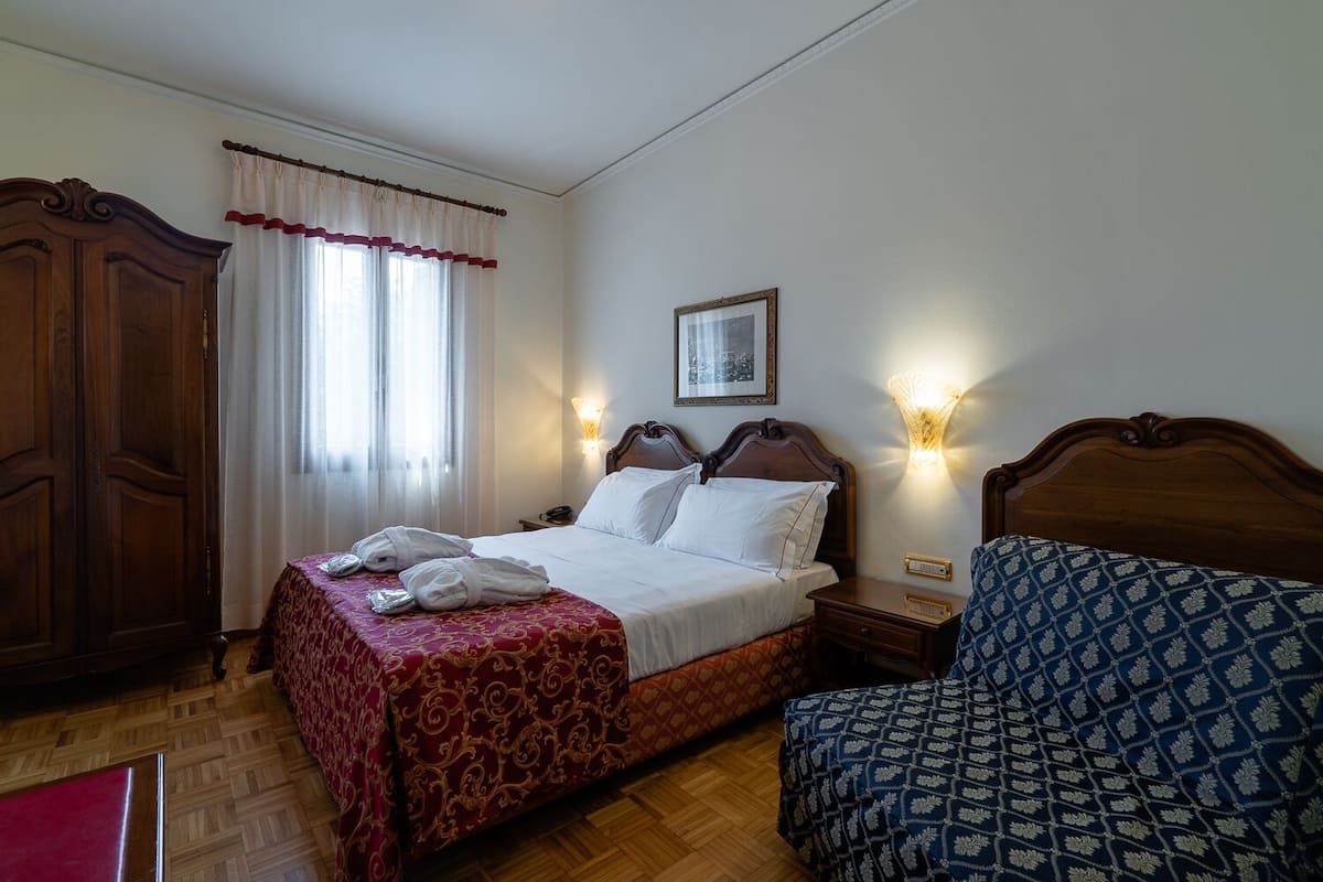 A hotel room with a wooden bed, a wooden wardrobe, a bedside table with a lamp, and a patterned armchair.