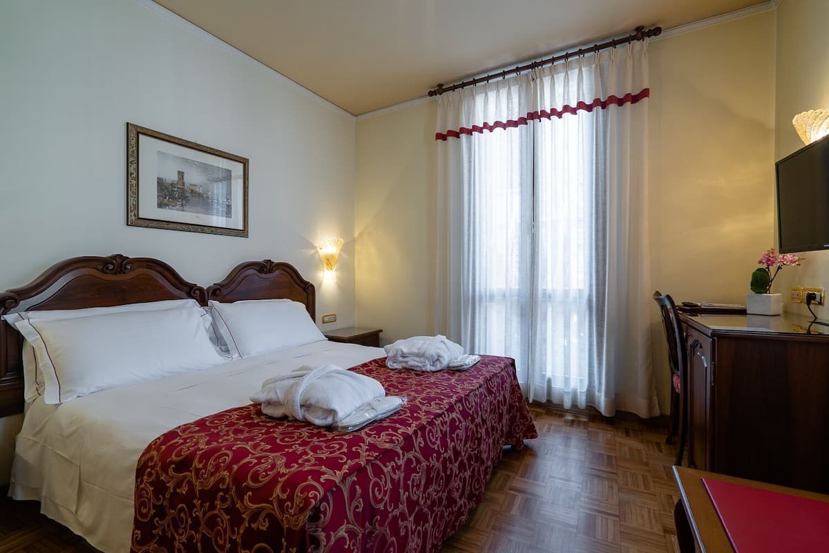 A hotel room with a double bed, two bedside tables, a desk with a chair, a lamp, a picture on the wall, and a window with curtains.