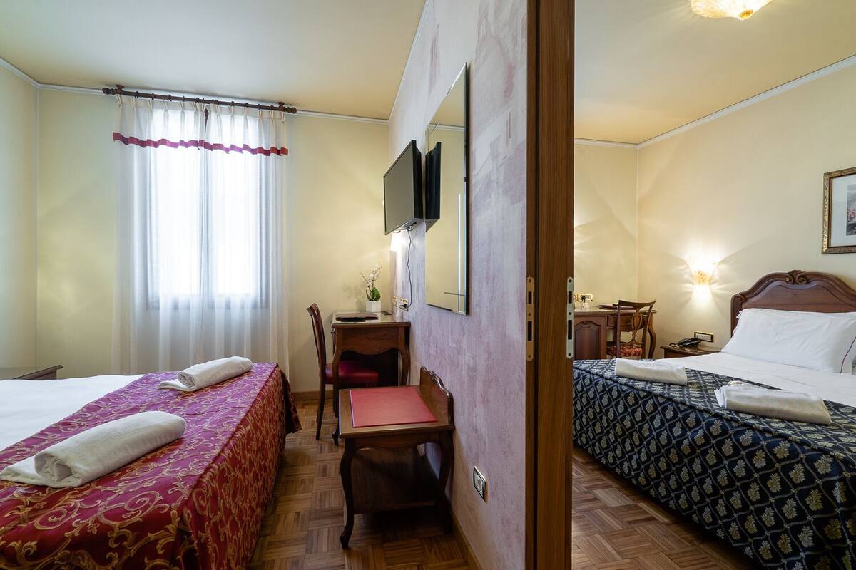 A hotel room with two beds, a desk, a chair, a TV, and a bedside table with a lamp.