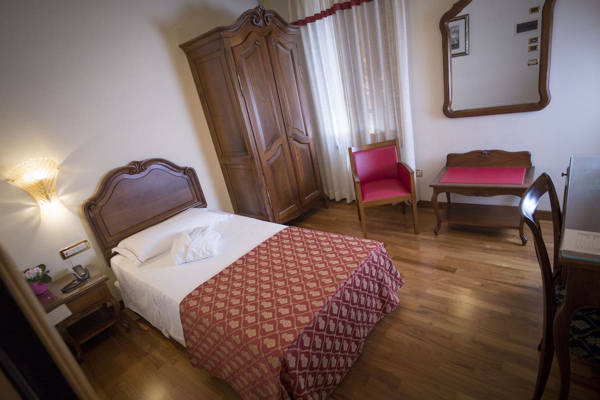 A neatly arranged bedroom with a bed, a wooden wardrobe, a desk with a chair, and a mirror on the wall.