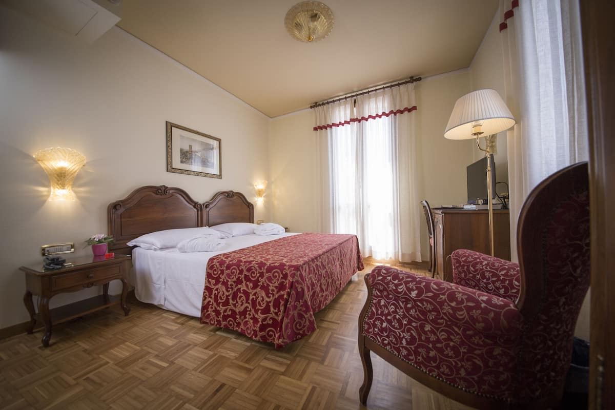 A hotel room with a wooden floor, a double bed with a red and gold bedspread, a nightstand with a lamp, a chair, and a mirror.