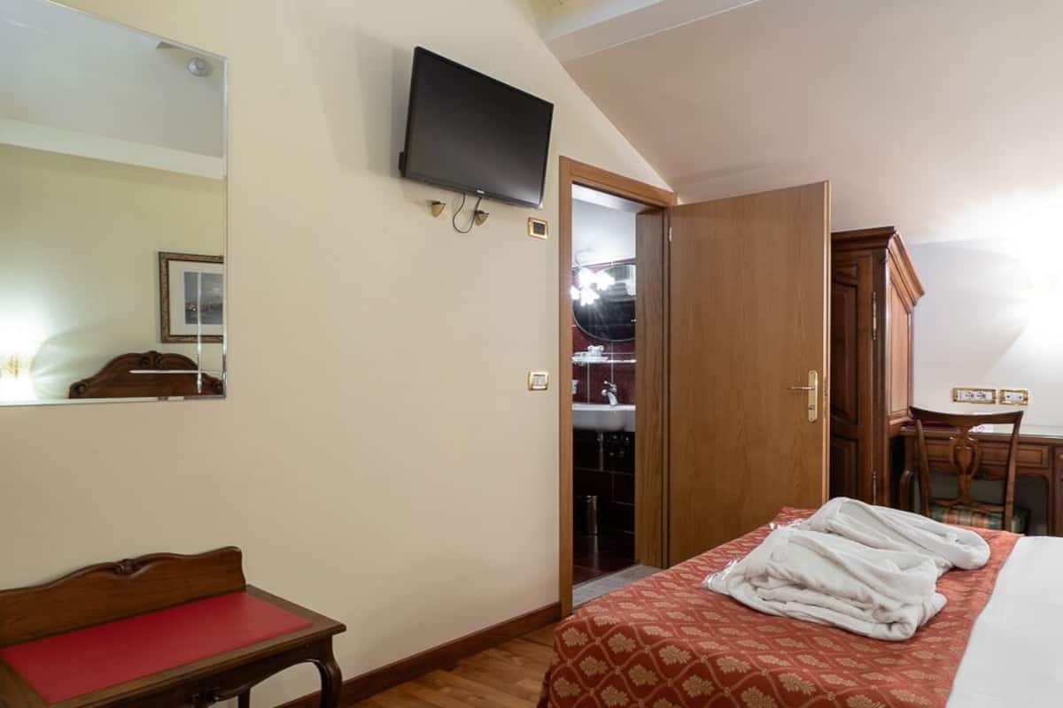 A hotel room with a wooden floor, a bed with a red patterned bedspread, a wooden desk with a red chair, a television mounted on the wall, and a wardrobe.