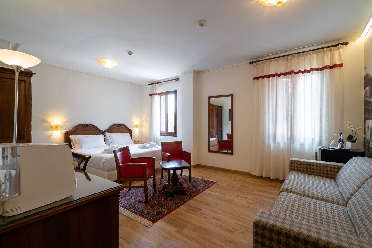 A hotel room with a bed, two chairs, a sofa, a desk with a lamp, a mirror, and a window with curtains.