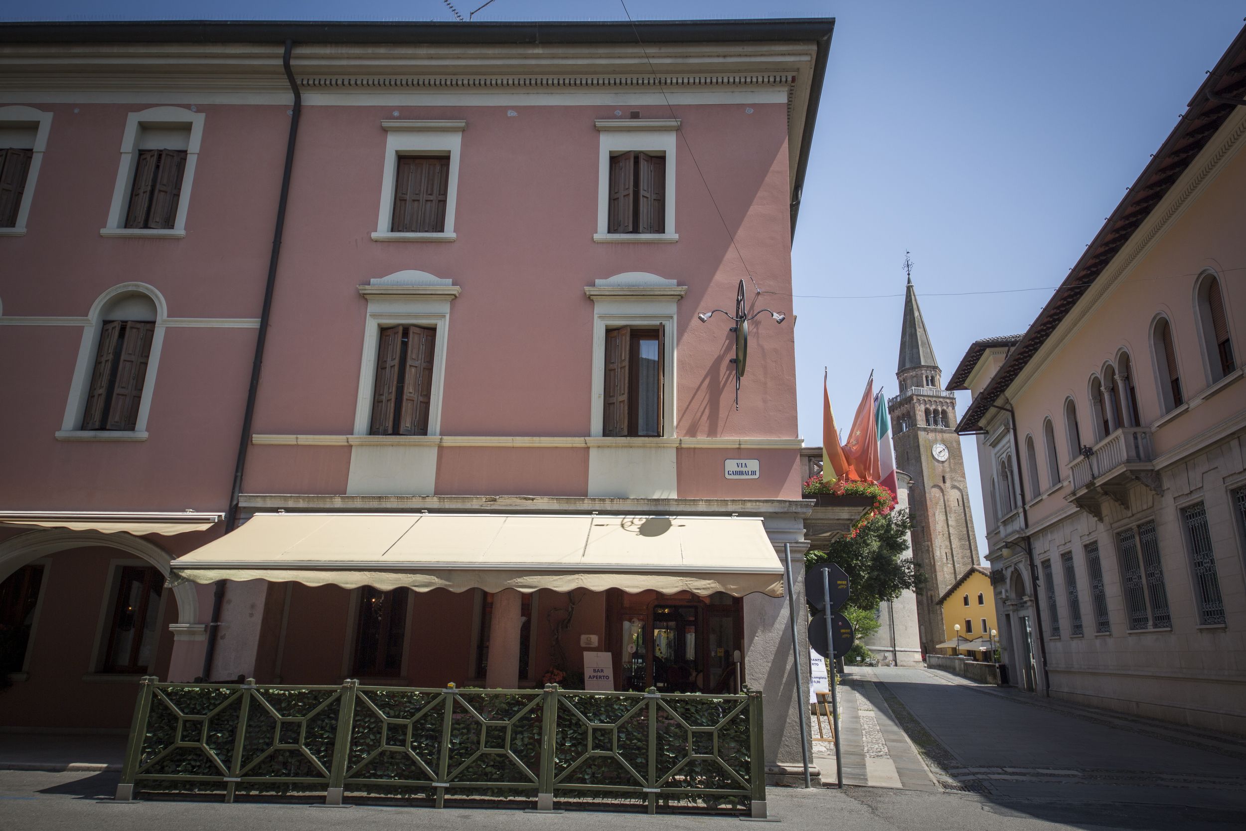 A pink building with a cafe on the ground floor, a clock tower in the background, and a sign reading "VIA CAMELLA".