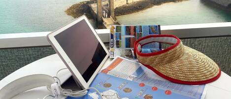 A tablet, headphones, a straw hat, and a map on a table with a view of the sea and a fort in the background.