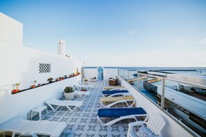 A sunlit outdoor deck with white lounge chairs and a patterned floor, overlooking a calm sea and a white building with a tower. - Hotel Miramar (Arrecife)