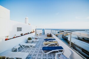 Terraza exterior iluminada por el sol con sillones blancos y suelo estampado, con vista a un mar tranquilo y un edificio blanco con una torre.
