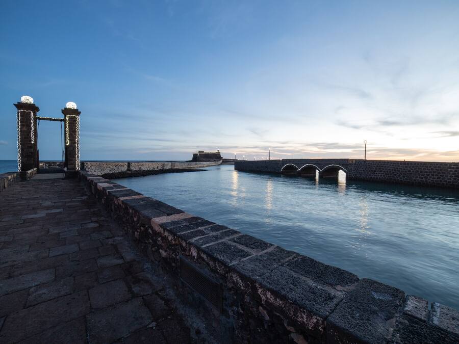 Un camino de piedra que lleva a un frente marítimo, con un muro de piedra y dos torres iluminadas.