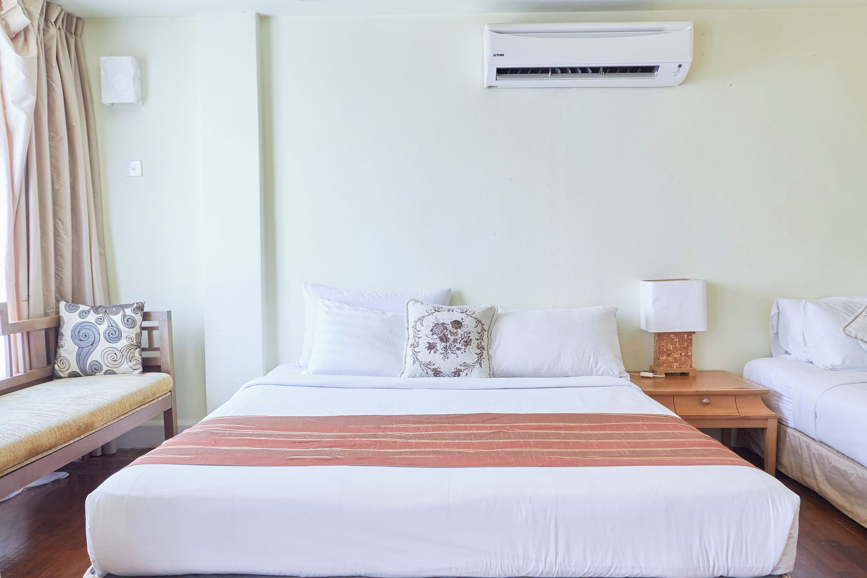 A hotel room with a bed, bedside tables, a chair, an air conditioning unit, and a window with curtains.