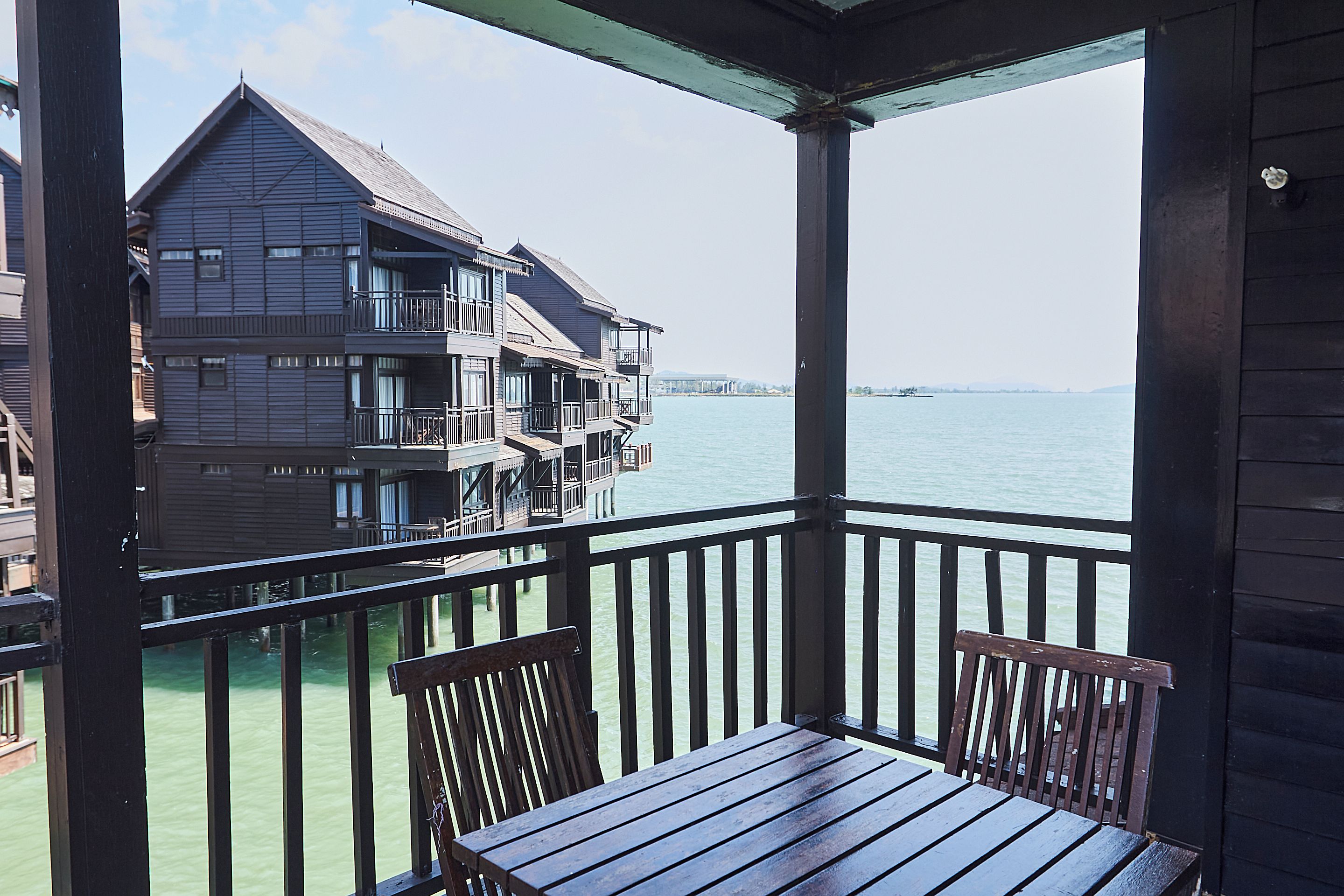 A wooden balcony with a table and chairs overlooking a body of water and a multi-story building.