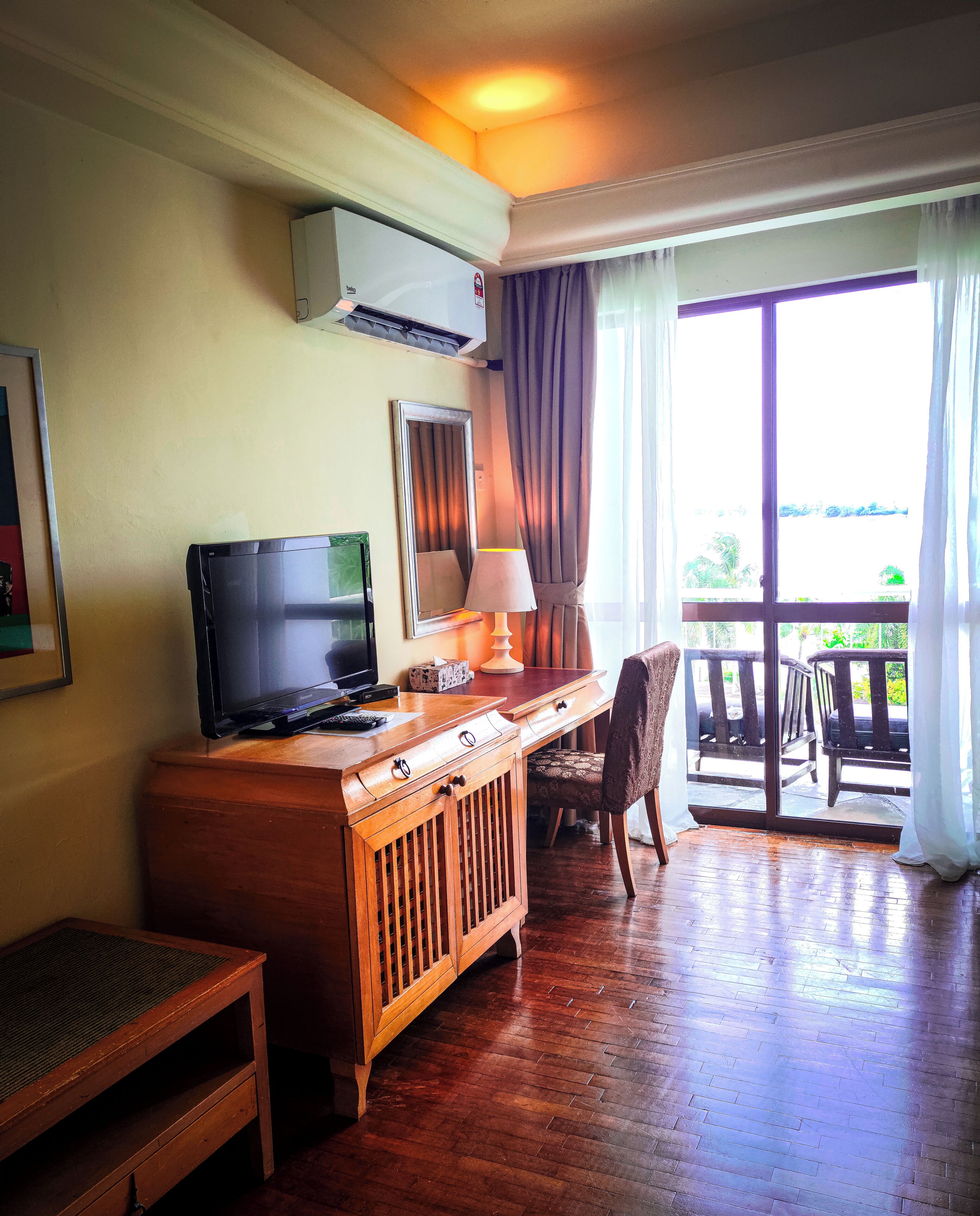 A room with a wooden desk, a television, a lamp, and a chair. There is a window with curtains and a view of the outdoors.