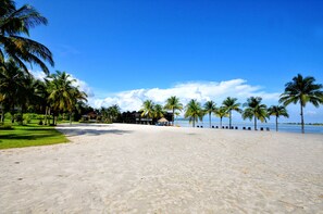 A beach with palm trees, a clear sky, and a building with a thatched roof. - Langkawi Lagoon Resort (Langkawi)