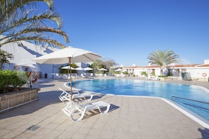 A swimming pool with lounge chairs and umbrellas, surrounded by palm trees and a building with a tiled roof. - Apartamentos Malibu Park (Adeje)